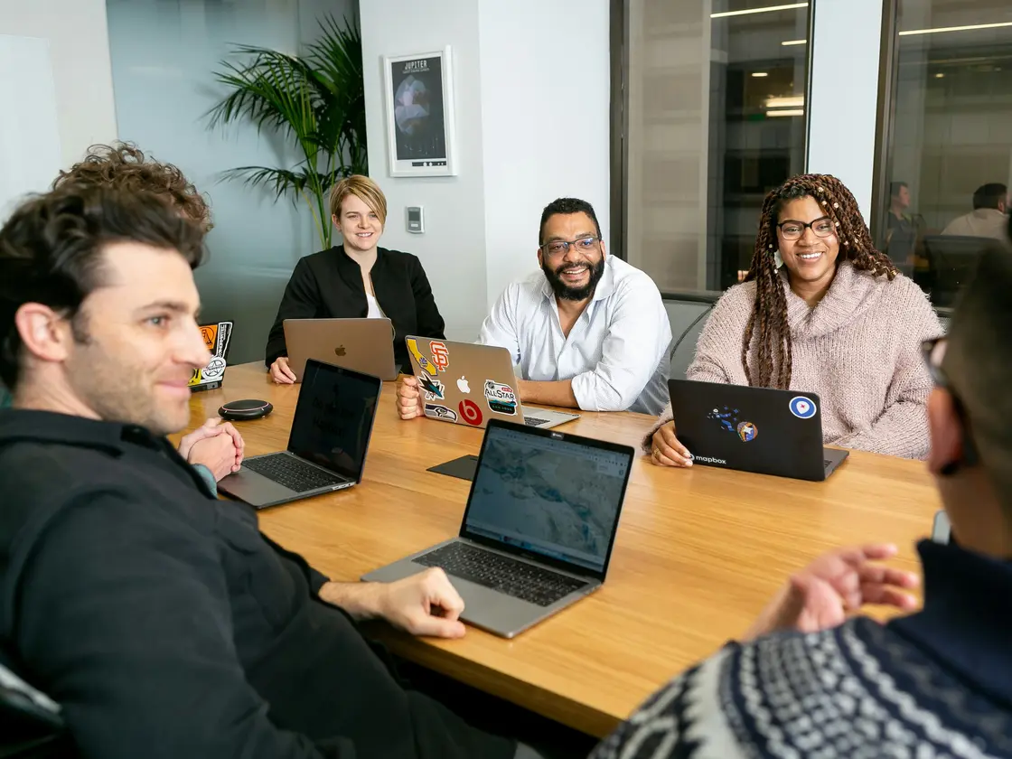 A small team around a table with their laptops, listening as someone talks them through a plan
