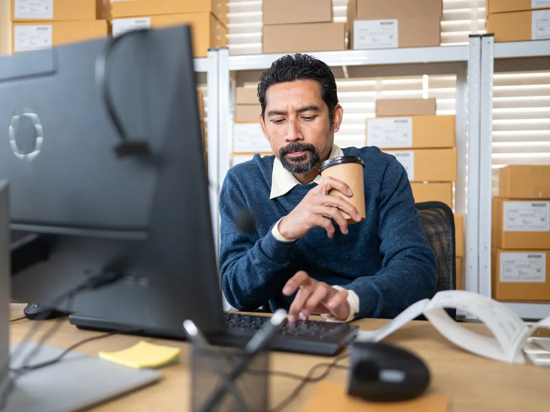 A business owner at his desk in the warehouse office, working with a coffee in hand