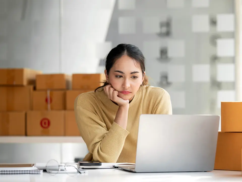 A business owner among boxes of stock, frowning at a new system on her laptop
